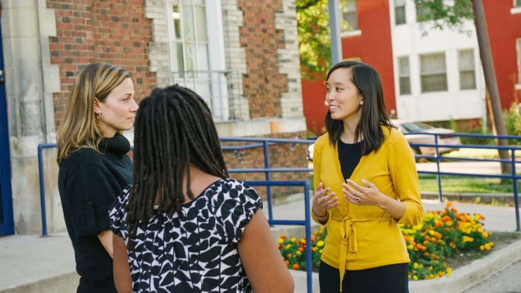 a woman wearing yellow speaks with two other women on a sidewalk