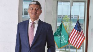 a Black man in a suit and tie smiling in front of the American flag and a Wayne State University flag