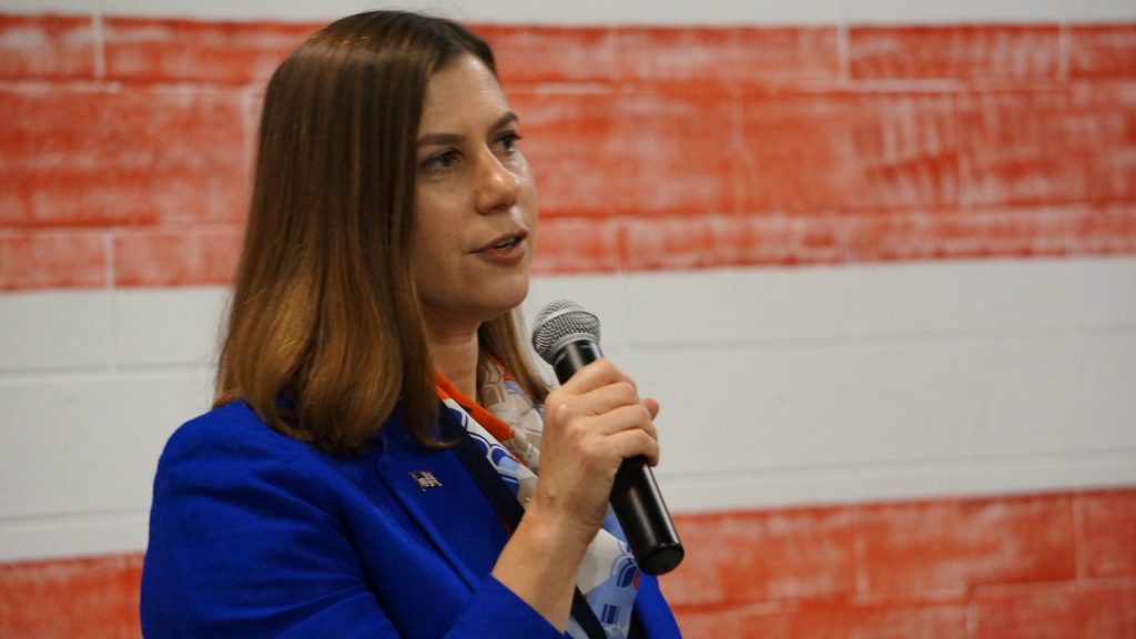 a woman in a blue blazer speaks into a microphone in front of an American flag