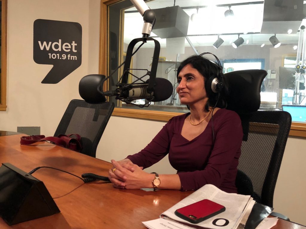 Shikha Dalmia sits at a table and speaks into a microphone in front of the WDET logo
