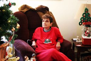 an elderly woman sits in a recliner, wearing a red sweater with Christmas tree embroidery