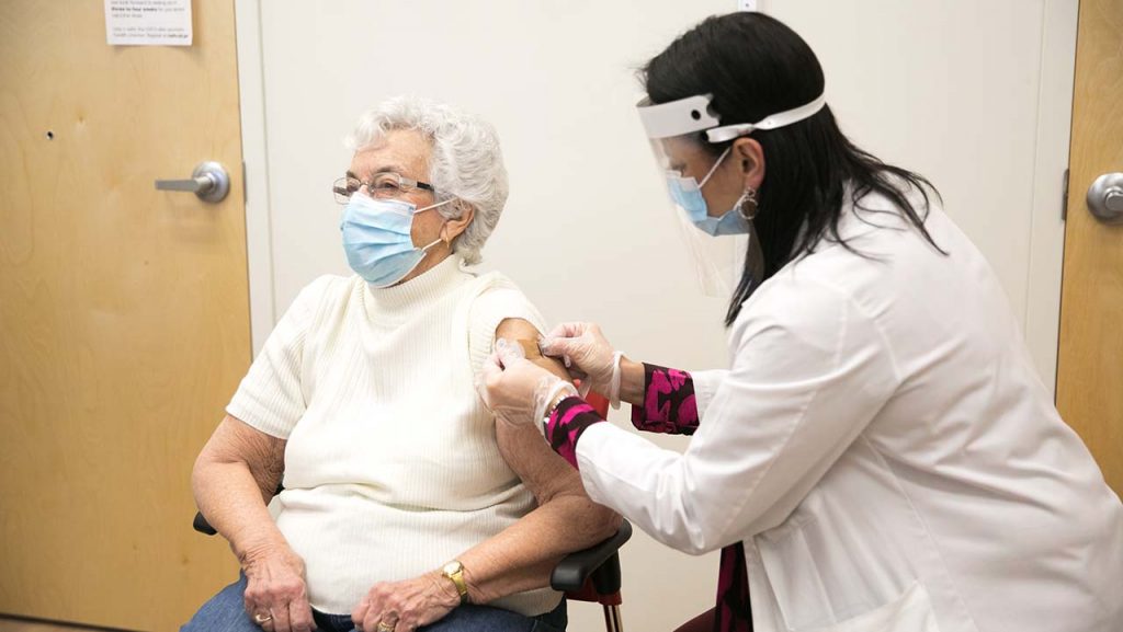 an older woman wearing a surgical mask receives a vaccination from a younger woman wearing a surgical mask, face shield and a lab coat