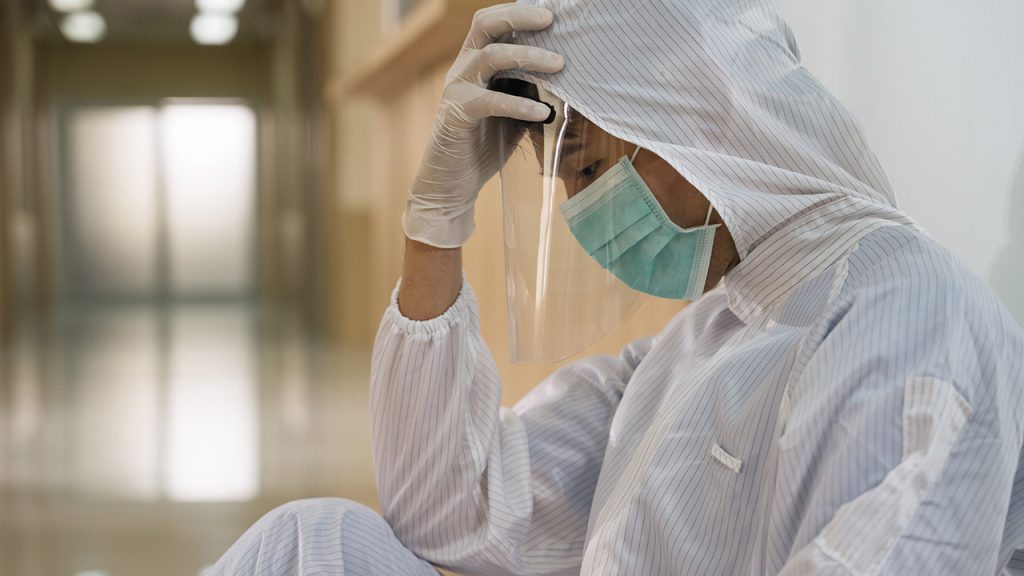 A doctor in a protective suit and mask sits on the floor looking tired.