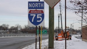 The blue and white I-75 sign is seen on the street with snow on the ground and orange barrels in the background.