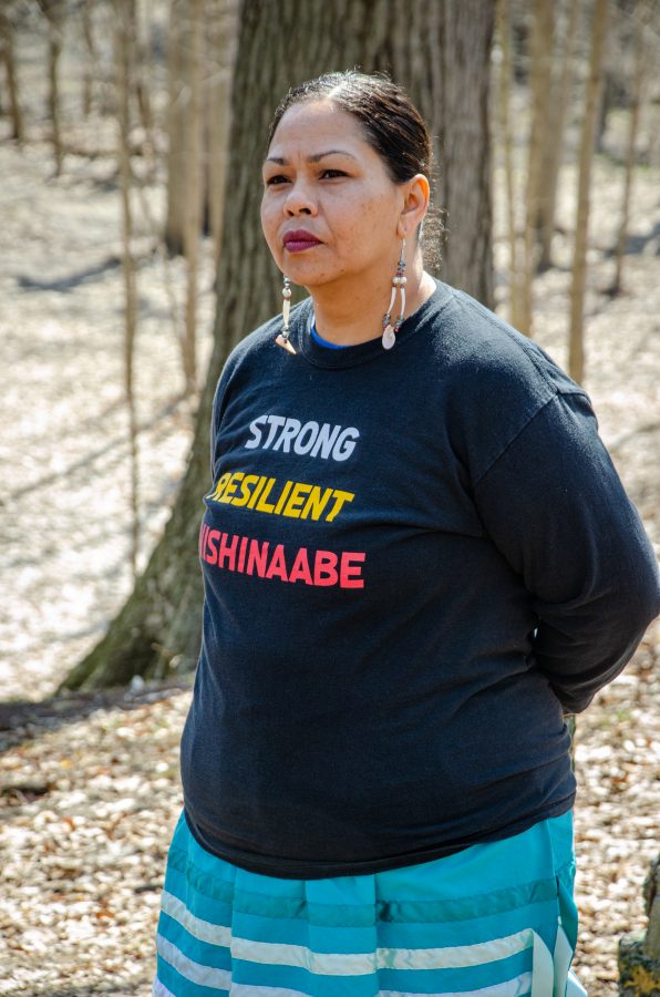 Indigenous woman stands in a forest with a shirt that reads "strong, resilient, Waawiyaataanong"