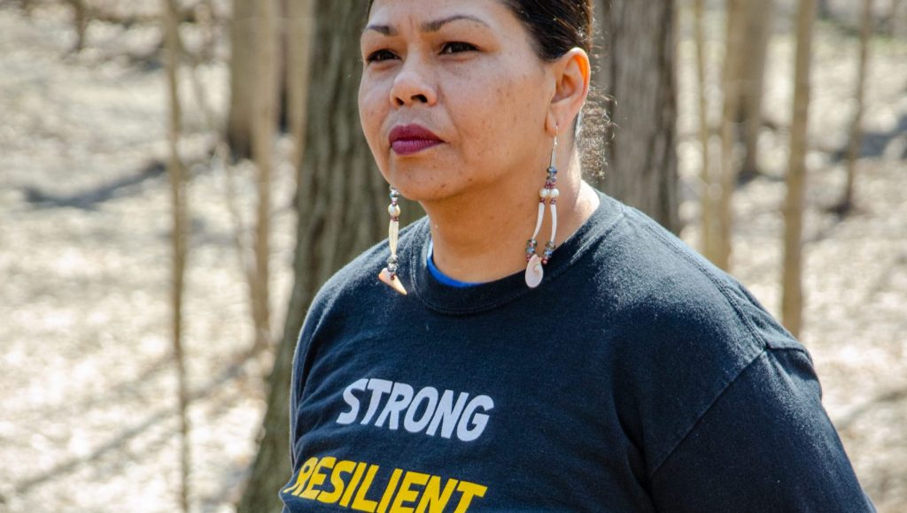 Indigenous woman stands in a forest with a shirt that reads "strong, resilient, Waawiyaataanong"
