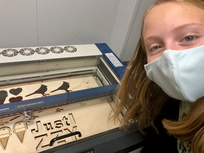 a teenage girl wearing a protective face mask smiles in front of a machine with wooden cutouts of figures like birds, hearts and ice cream cones