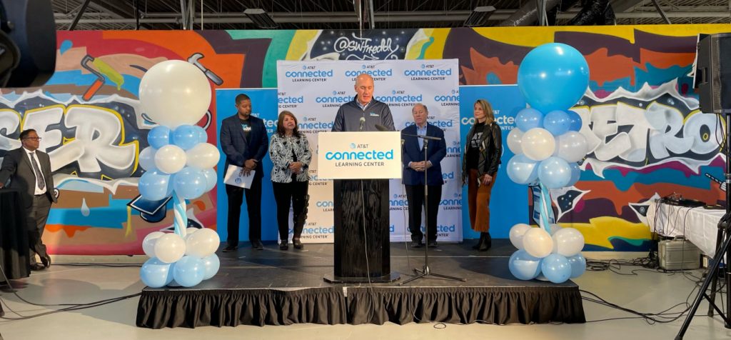 John Stankey stands at a podium on a stage decorated with blue and white balloons alongside Eva Garza Dewaelsche and Mayor Mike Duggan
