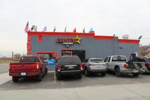 Image shows outside of Coney Star Halal, a grey building with red trim and flags on top. There are cars parked in the parking lot.