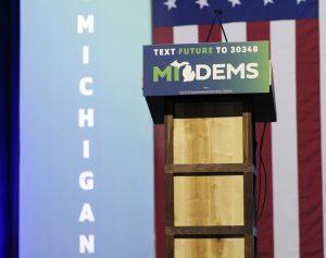 podium that reads "MI Dems" in front of an American flag and a blue sign that reads "Michigan"