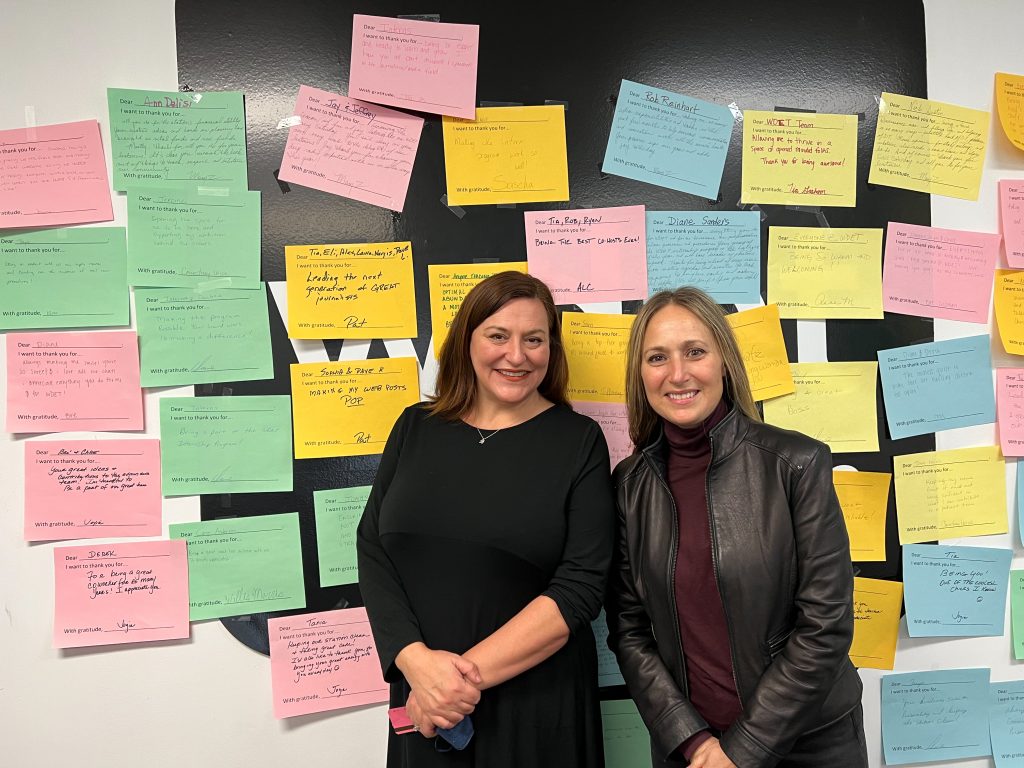 Courtney Burkett and Heather Raffo smile in front of the WDET logo (which is covered with multi-colored notes of appreciation from WDET staff) at our studios