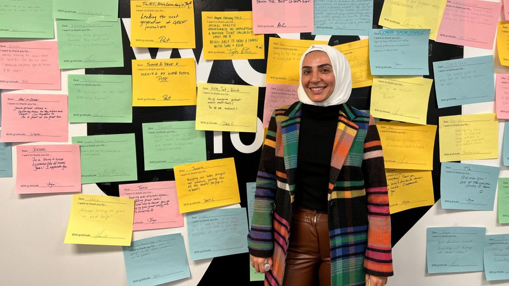 nawal alsaeed smiles in front of the WDET logo (which is covered with multi-colored notes of appreciation from WDET staff) at our studios