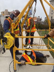 men in orange vests and hard helmets stand on a sidewalk, around a yellow metal structure with cables hanging from it. One man is partially submerged into a manhole in the sidewalk.