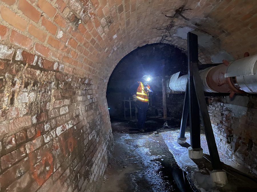 a person wearing an orange vest and a headlamp stands at the other end of a narrow brick tunnel with a pipe running through it