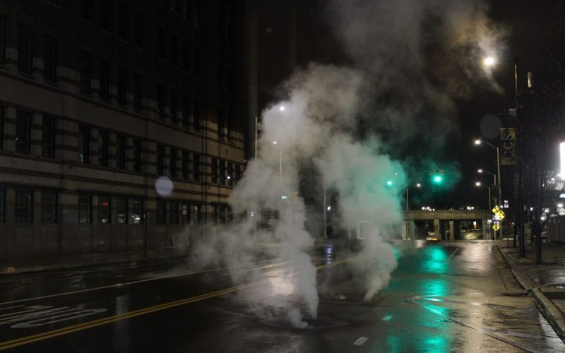 A plume of white steam rises out of a manhole cover on a Detroit street at night.