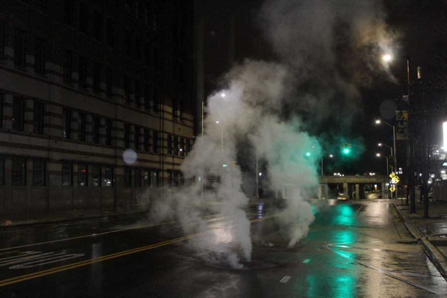 A plume of white steam rises out of a manhole cover on a Detroit street at night.