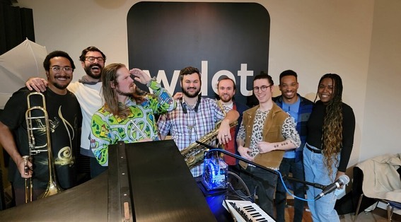 seven guys pose with various instruments in front of the WDET logo with CultureShift's Tia Graham