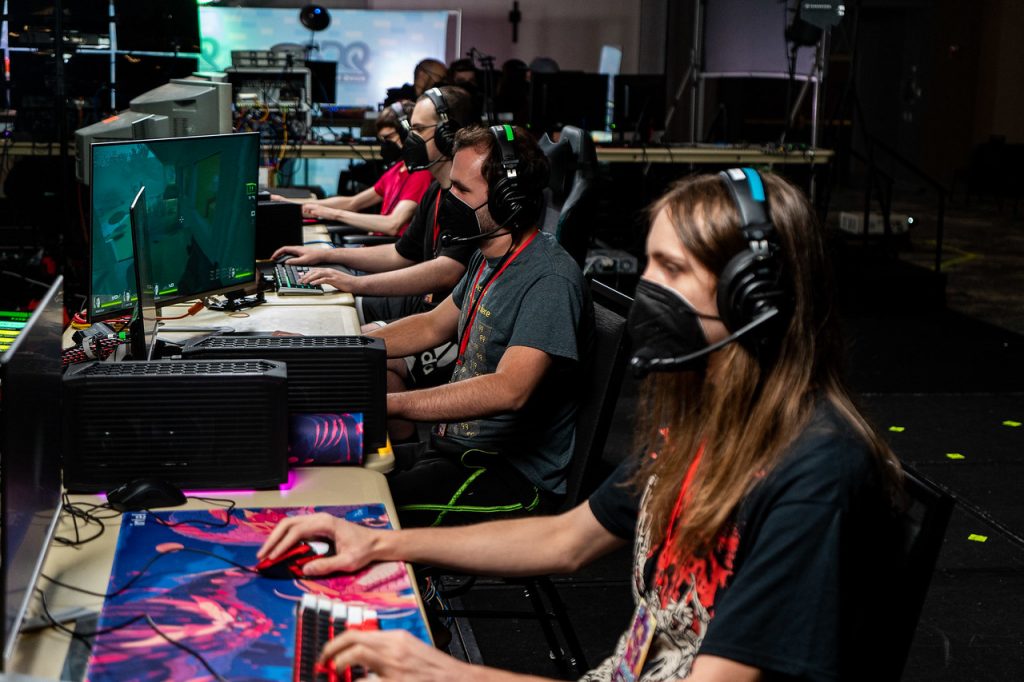 people sit in a row wearing headsets and face masks, playing video games