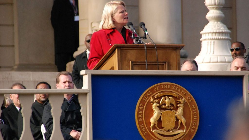 a woman in a red coat speaks at a podium