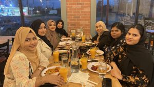 Seven young women with hijabs smile at a table with plates of food and drinks