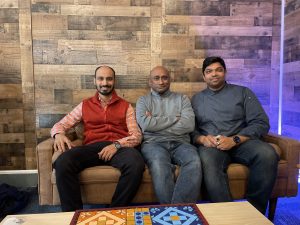 three Bangladeshi men smile on a couch against a brick wall