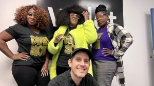 three Black women pose in front of the WDET logo with a white man