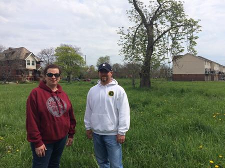 two adults in hoodies stand in a grassy field with houses in the distance