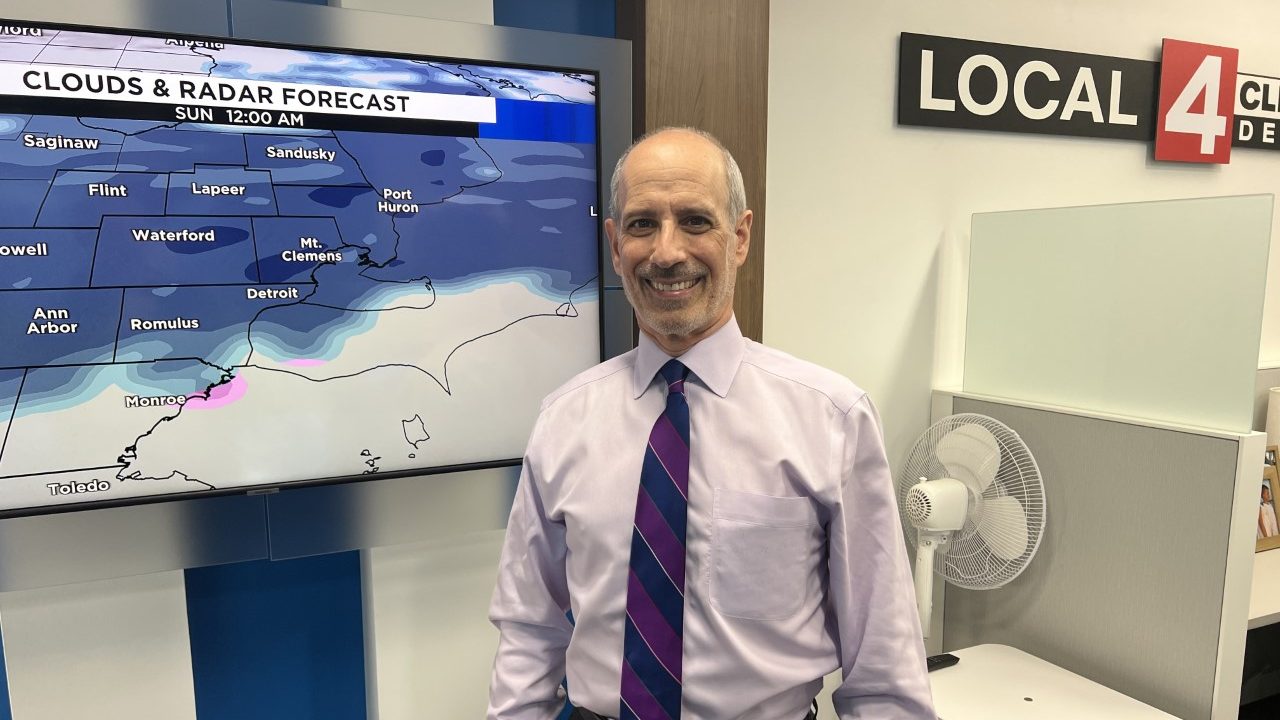 white man with a purple shirt and tie smiles in front of a weather map