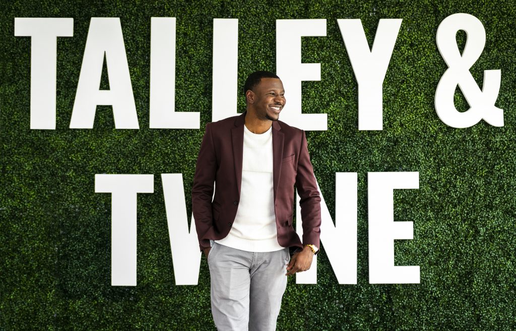 a Black man in a blazer smiles in front of greenery with "Talley & Twine" sign