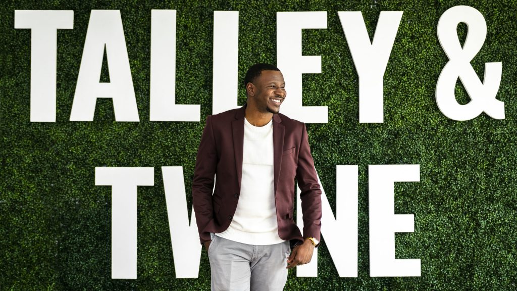 a Black man in a blazer smiles in front of greenery with "Talley & Twine" sign