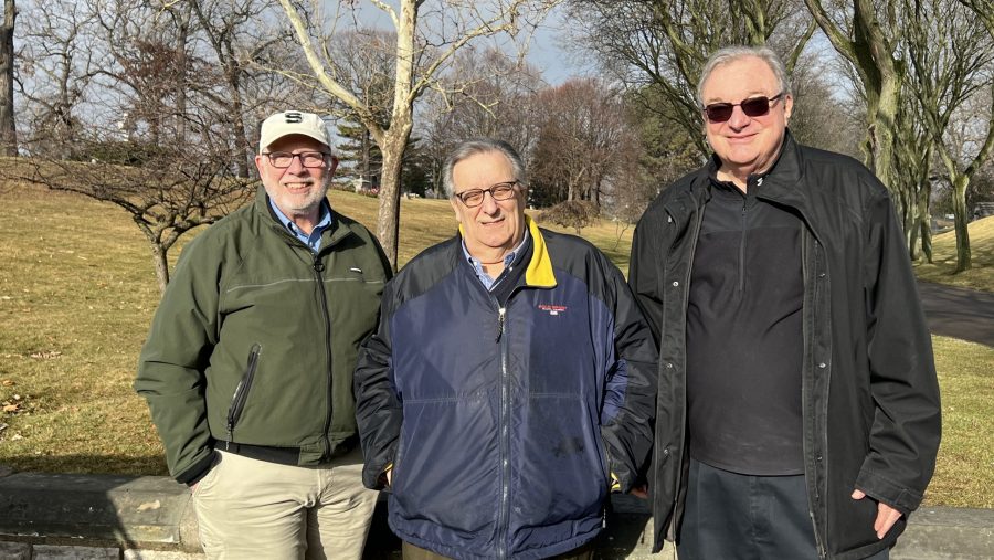 three men in winter jackets smile in a sunny, grassy area