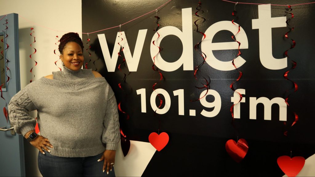 a Black woman in a stylish gray sweater poses in front of the WDET logo