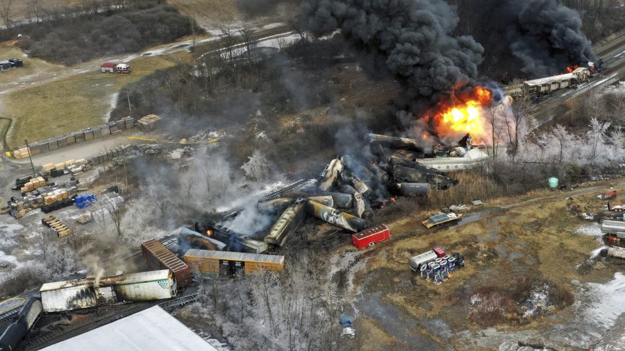 Overhead view of several train cars derailed from the tracks with one on fire. Dark smoke clouds the area