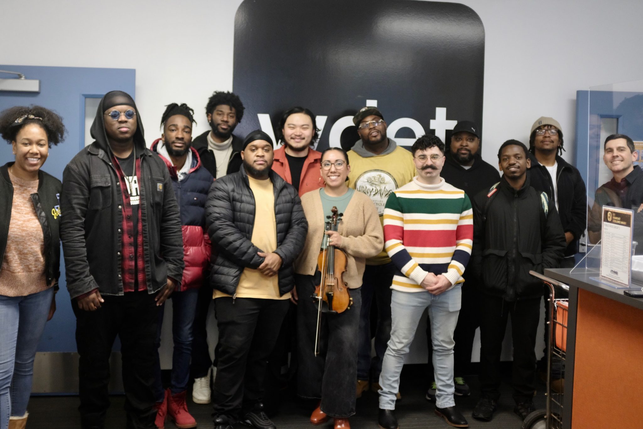 a group of twelve musicians pose in front of WDET's logo with Sam Beaubien. One of them is holding a violin