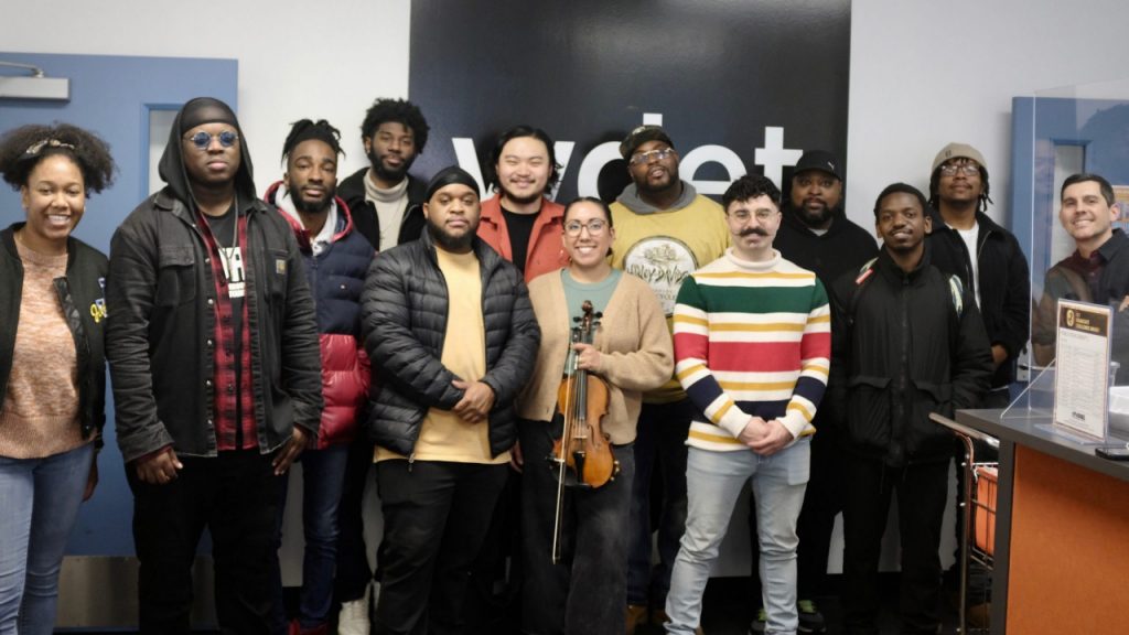 a group of twelve musicians pose in front of WDET's logo with Sam Beaubien. One of them is holding a violin