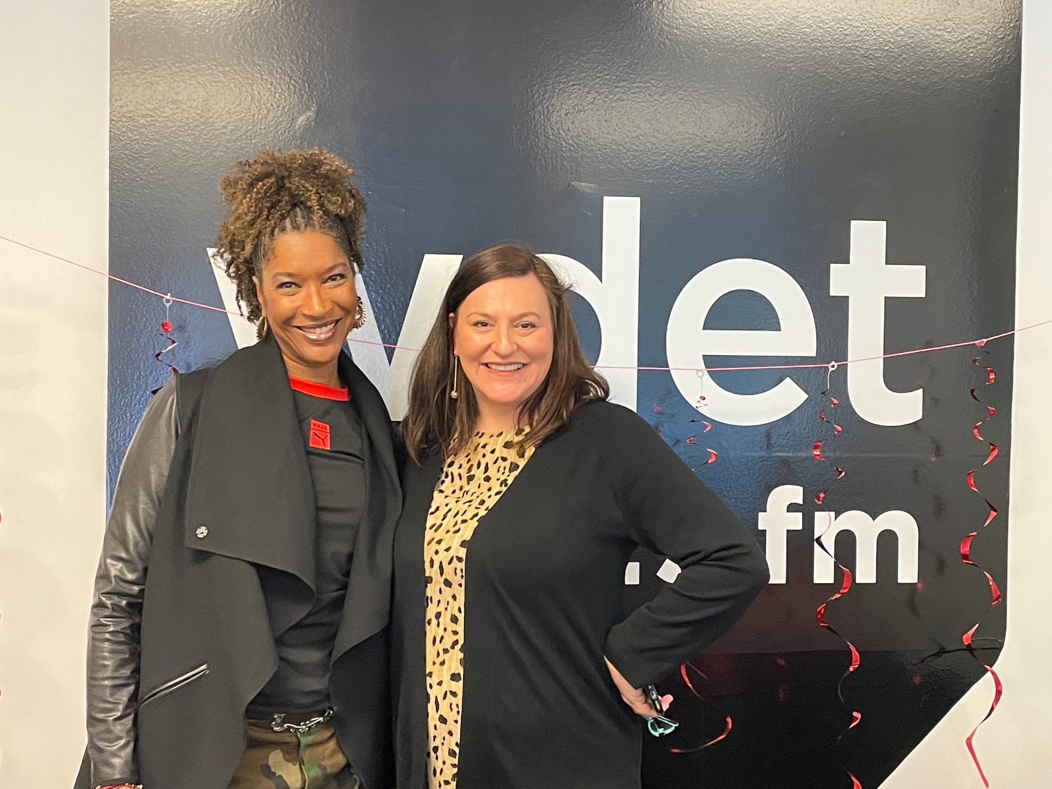 two women smile in front of the WDET logo decorated with red streamers for Valentine's Day