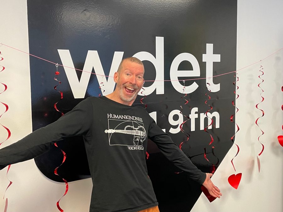 a white man smiles with his arms outstretched in front of the WDET logo