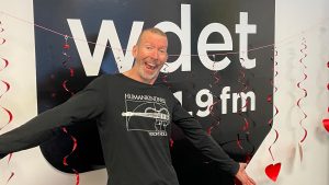 a white man smiles with his arms outstretched in front of the WDET logo