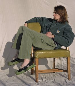 a person with shoulder-length hair poses in an oversized green outfit with the Carhartt patch during golden hour