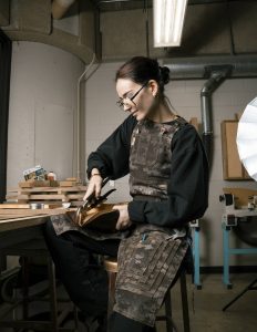 a person with their hair pulled back sits in a workshop wearing a canvas apron with several layered pockets