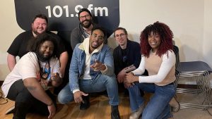 Five musicians crouch in front of the WDET logo in our live studio with CultureShift's Tia Graham