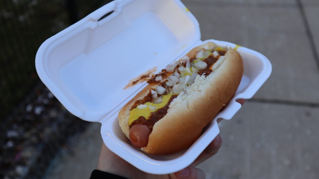 A hand holds a hot dog piled high with chili sauce, mustard and onions in a to-go box.