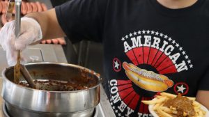 A person wearing an American Coney Island t-shirt and gloves scoops chili onto a plate of french fries