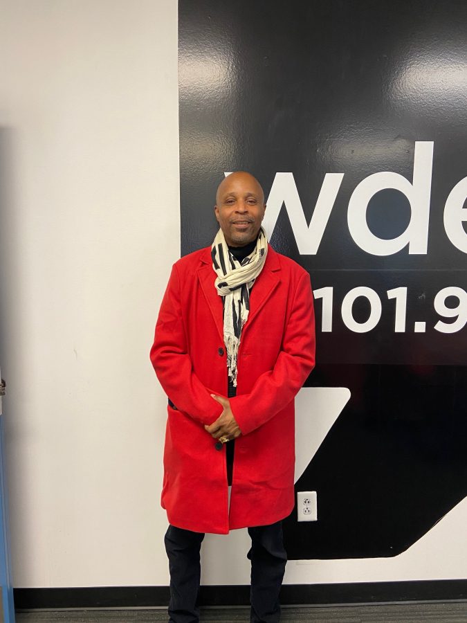 A Black man in a red coat and white scarf smiles in front of the WDET logo