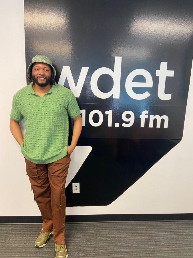 A young Black man in a green shirt and hat smiles in front of the WDET logo