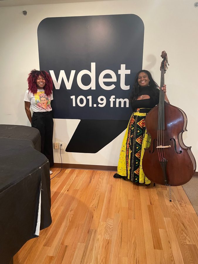Two Black women smile in front of the WDET logo, one of them holding an upright bass.