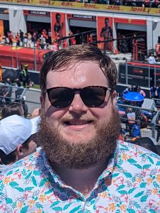 A white man with a beard, colorful shirt and sunglasses smiles in a crowd.