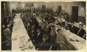 Black and white image of many men dressed in suits sitting at a dining table