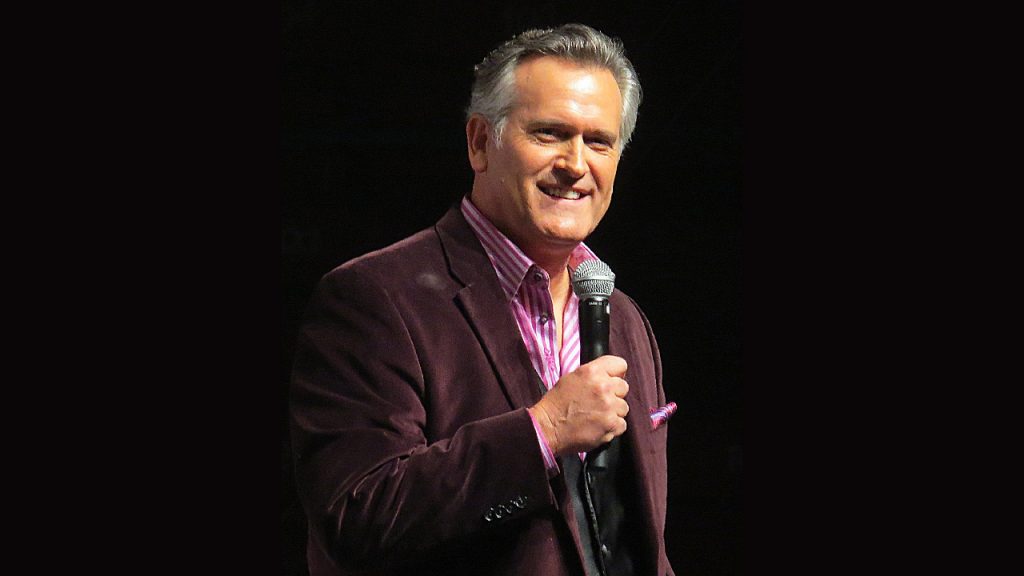 Actor Bruce Campbell stands onstage with a microphone wearing a suit