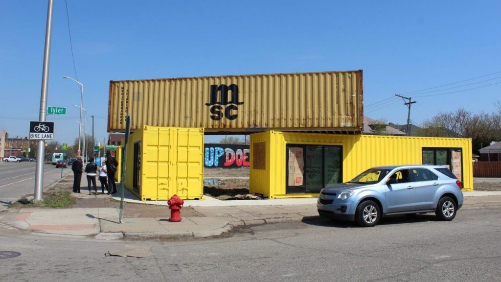 A shipping container is stacked on top of two bright yellow shipping containers. Behind them you can see part of a mural that says "What Up Doe."